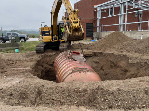 Excavator lifting a rusted underground storage tank out of the ground beside a roadway